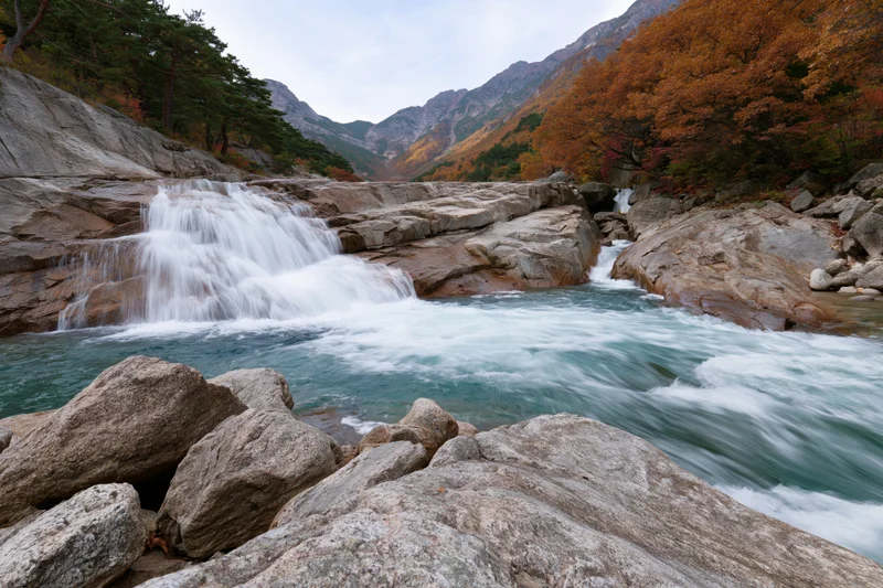 Waterfall cascading over granite ledges into a river surrounded by autumn forest in mountain valley