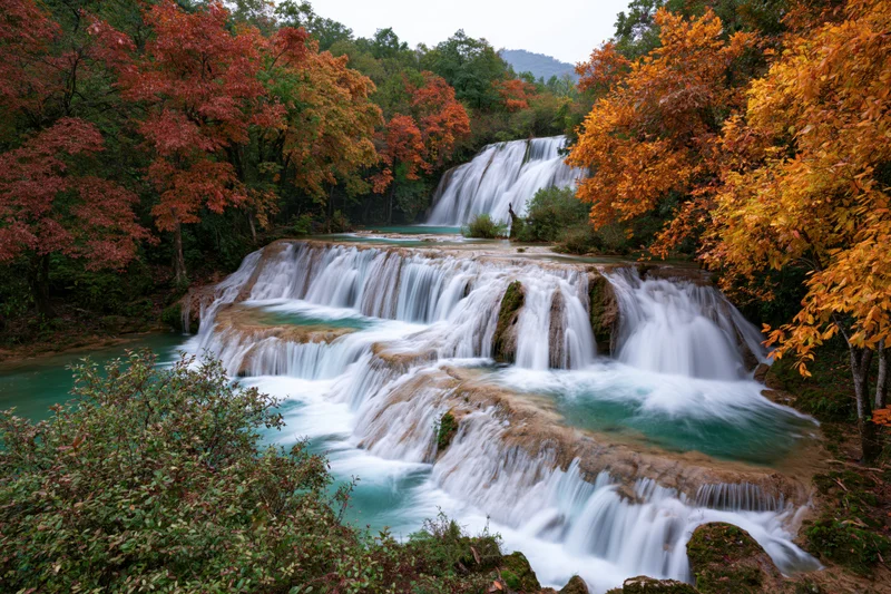 Multi-tiered waterfall cascading over limestone rocks surrounded by autumn-colored trees in a jungle landscape