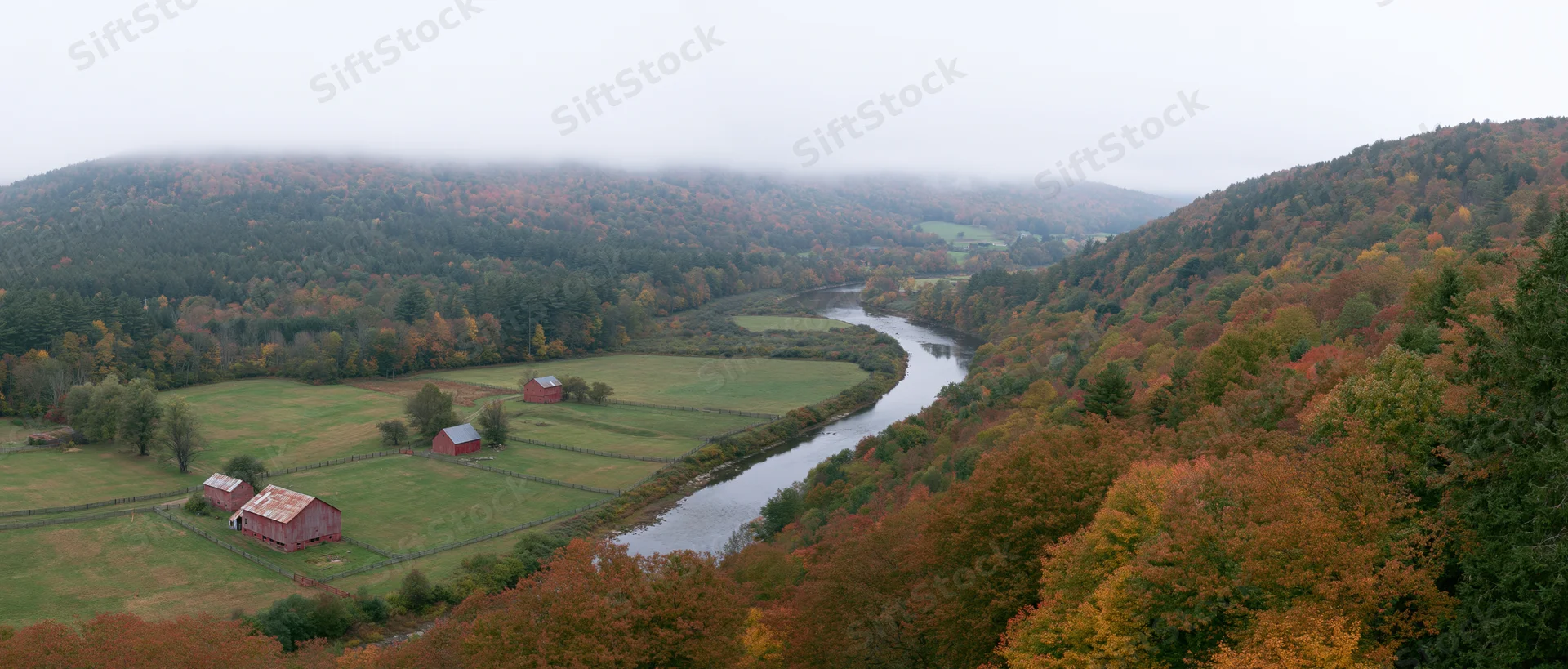 Winding river through autumn forested valley with red barns and foggy hills