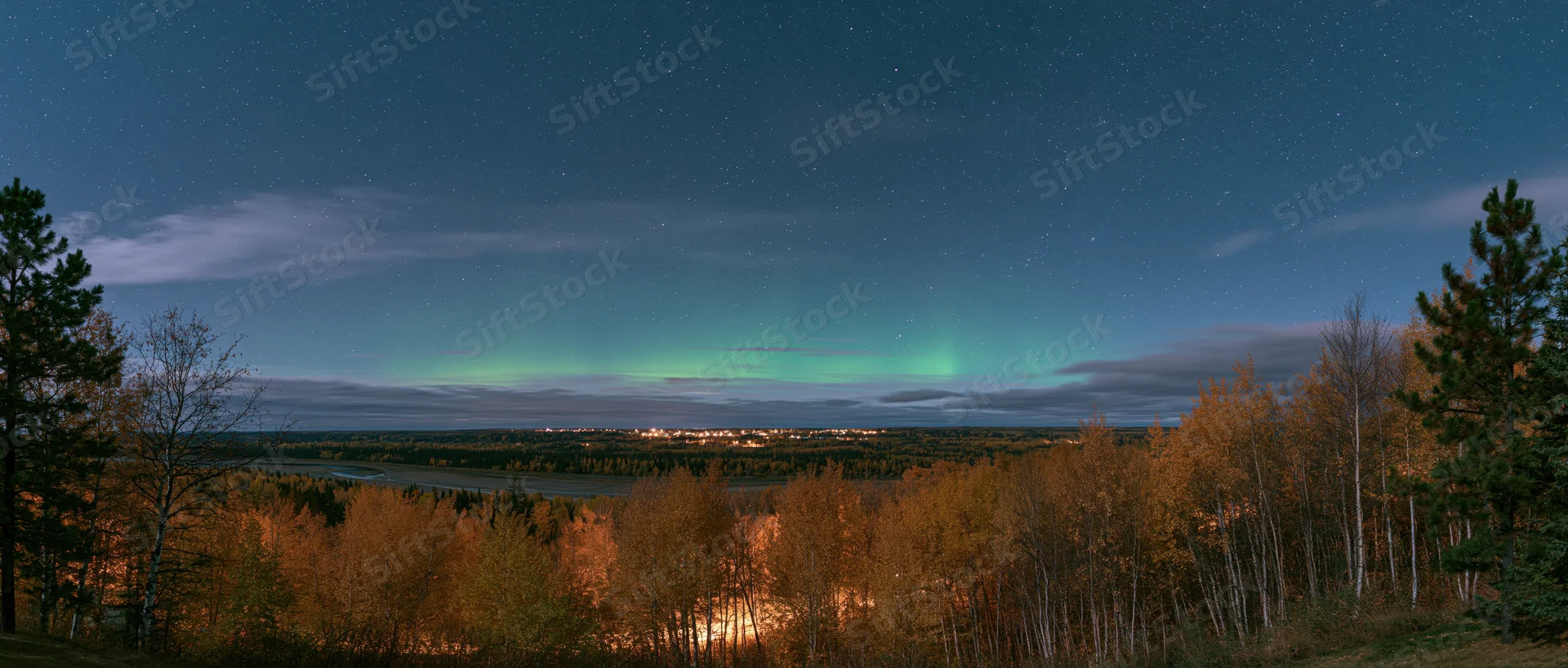 Aurora glowing over forested landscape with river and distant city lights at night