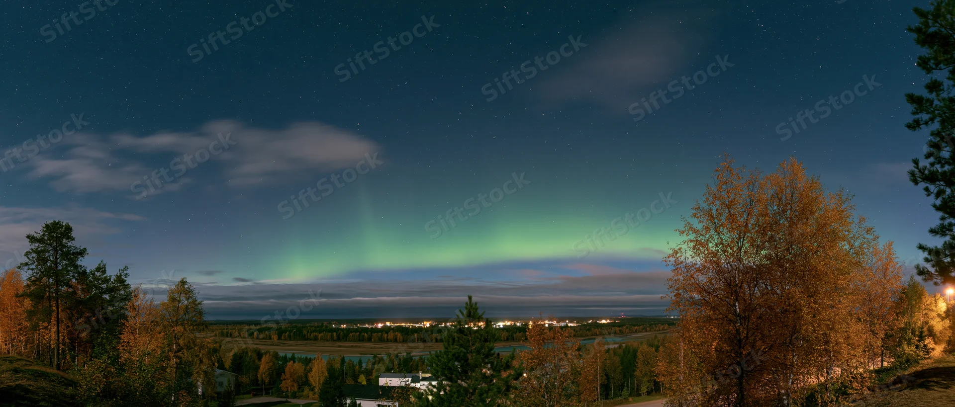 Aurora glowing green and teal low on the horizon over a forested landscape with autumn trees and distant town lights