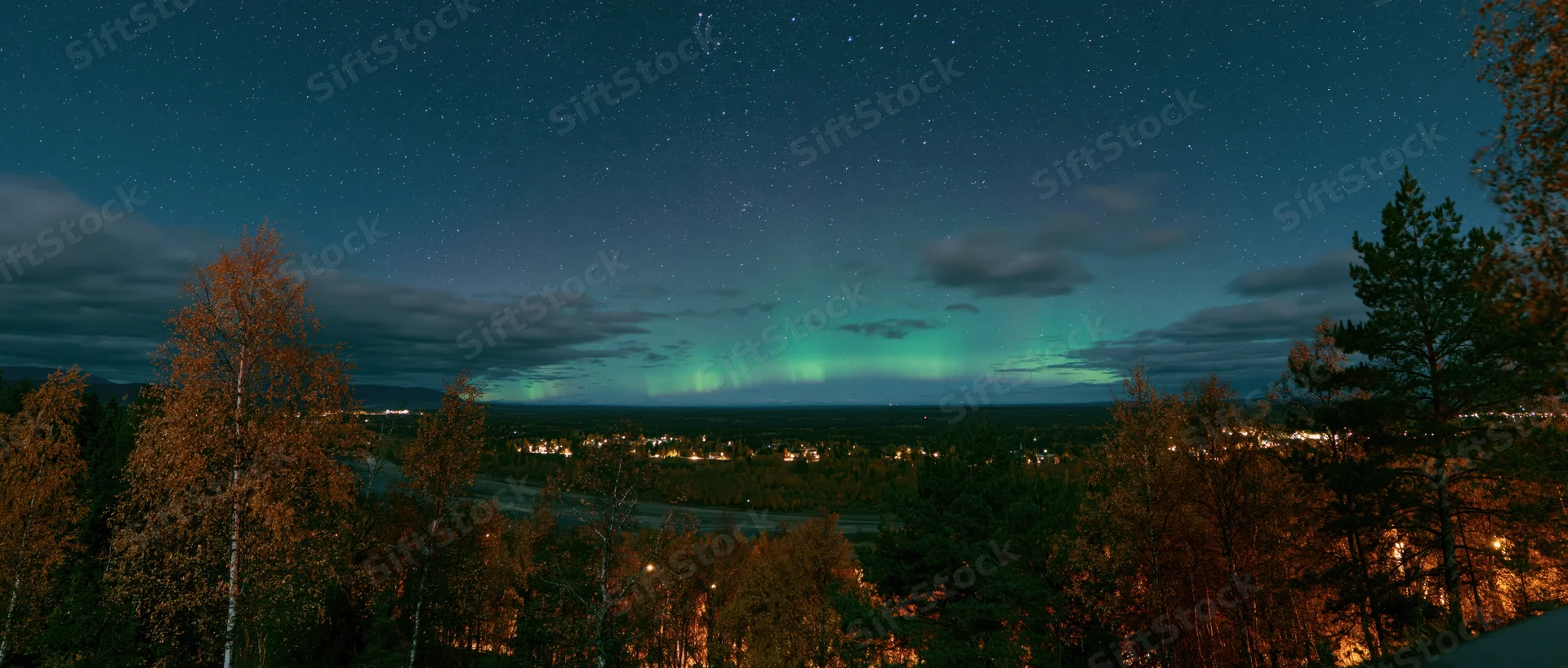 Aurora glowing green and teal over forested landscape at night with stars and distant town lights