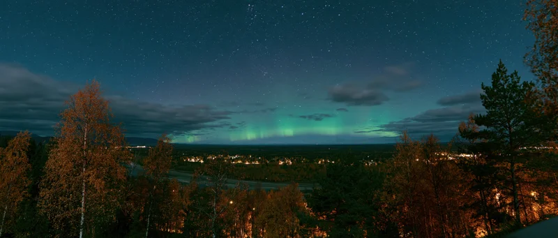 Aurora glowing green and teal over forested landscape at night with stars and distant town lights