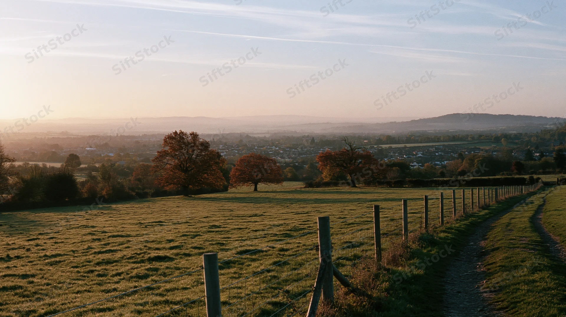 Wide panoramic view of a rural countryside field with a winding path and wooden fence at golden hour