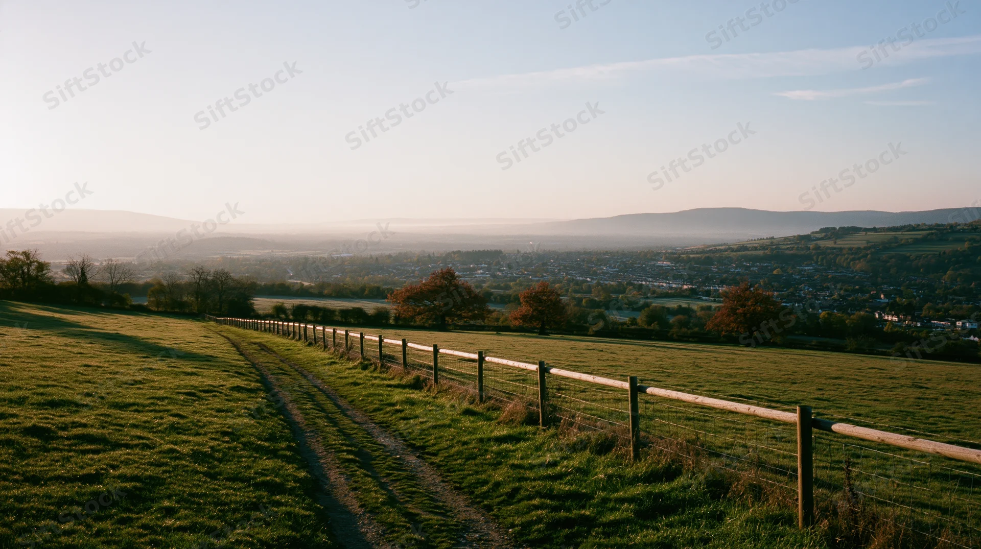 Wide panoramic view of open countryside with a winding path and wooden fence at golden hour