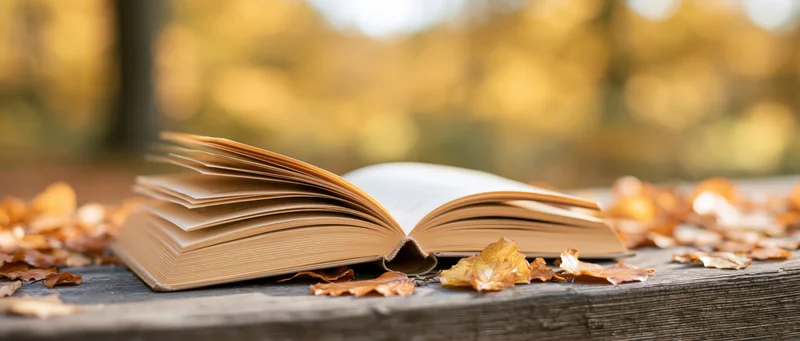 Open hardcover book resting on a wooden table surrounded by autumn leaves outdoors