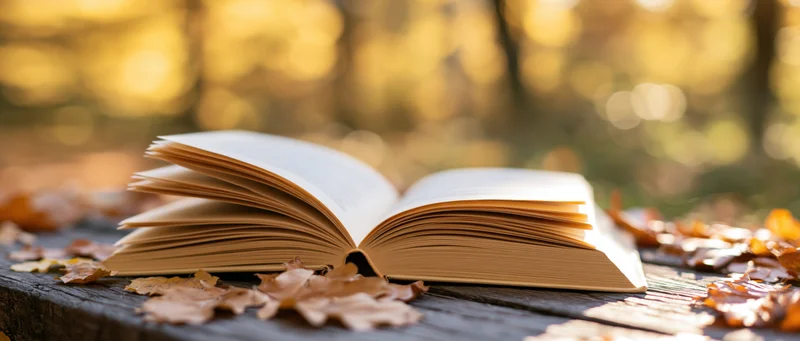 Open hardcover book with yellowed pages resting on a weathered wooden bench outdoors among autumn leaves