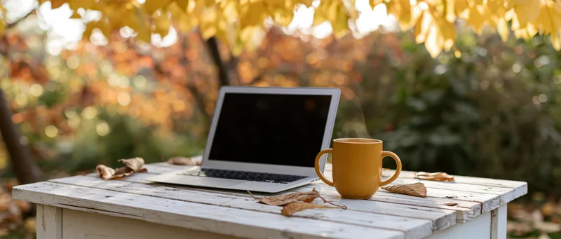 Open laptop with dark screen and steaming mug on white wooden table under autumn leaves