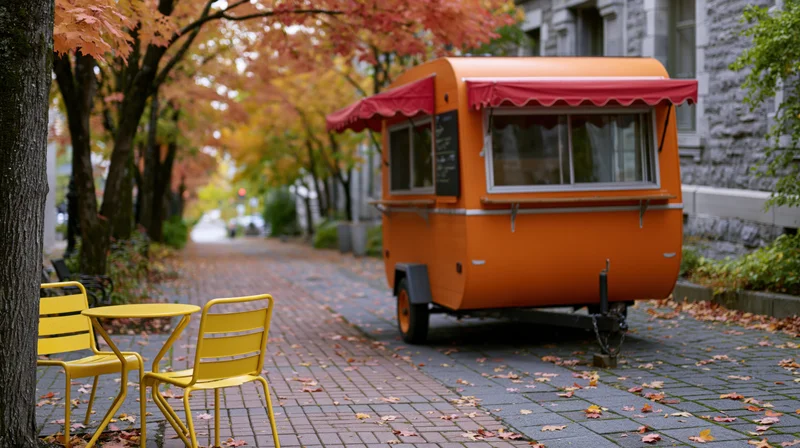 Orange food cart trailer with red awning parked on brick sidewalk lined with autumn trees, yellow chairs and table nearby