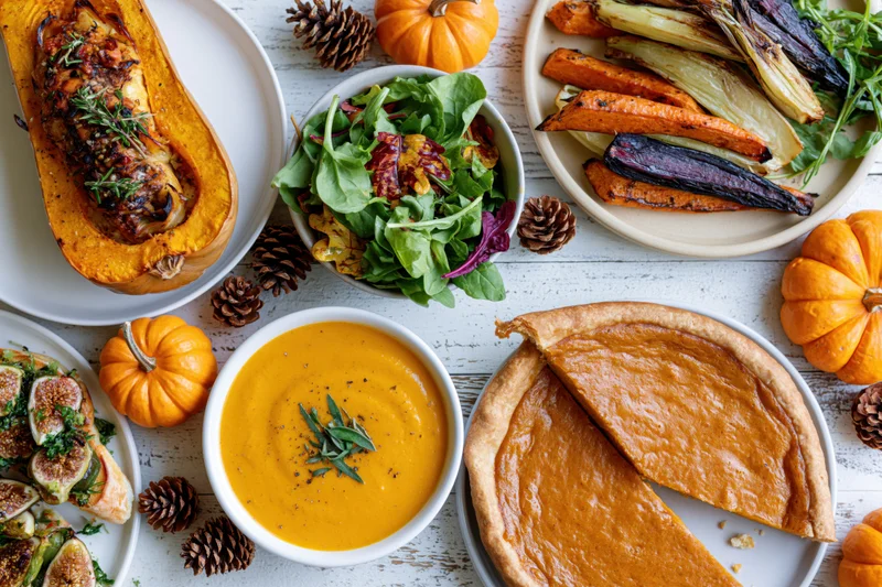 Overhead flat lay of a thanksgiving feast with pumpkin dishes, salad, pie, and autumn decor on a whitewashed wooden table