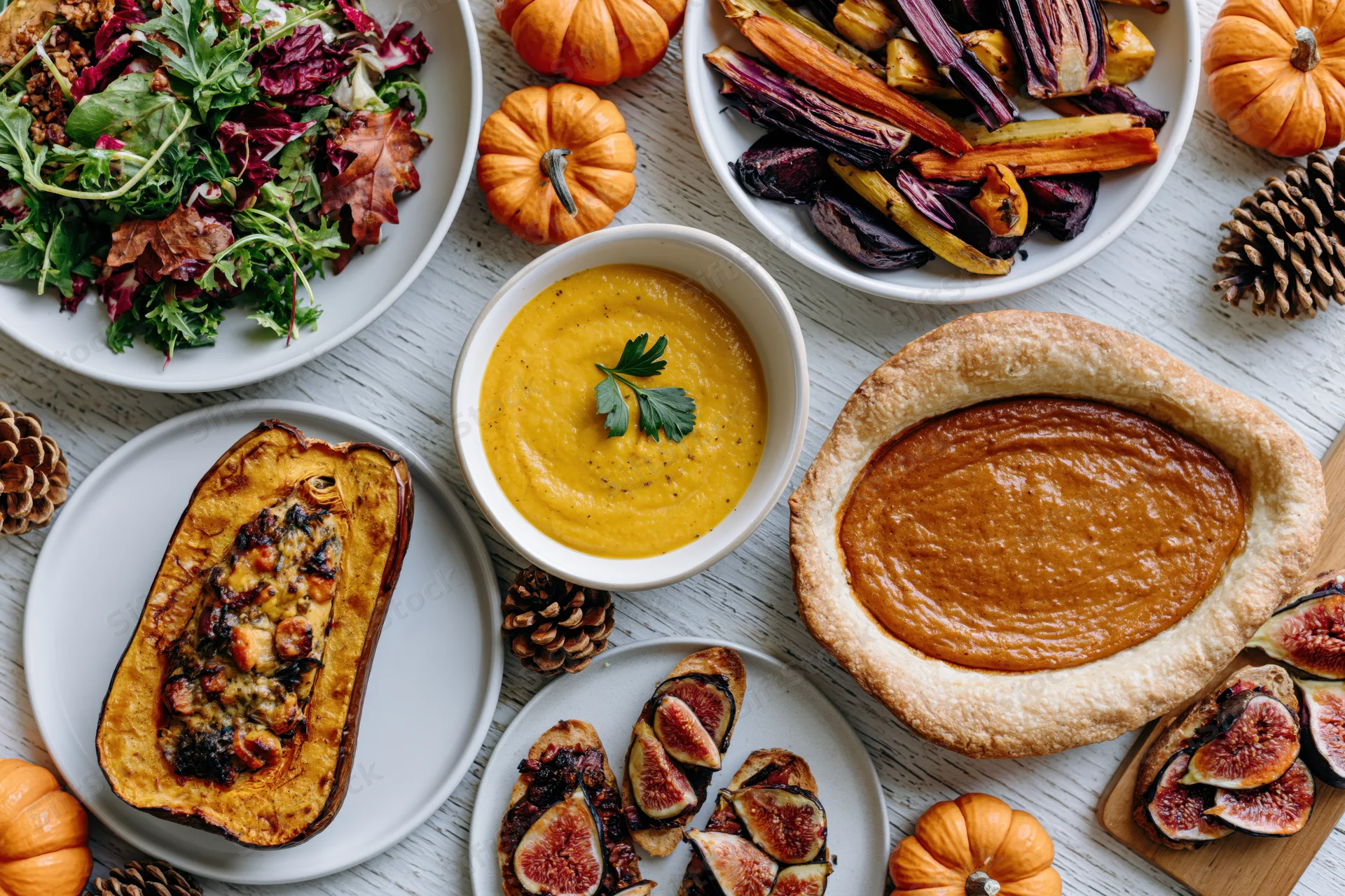 Overhead flat lay of thanksgiving dishes including pumpkin pie, roasted squash, soup, salad and figs on a whitewashed wooden table