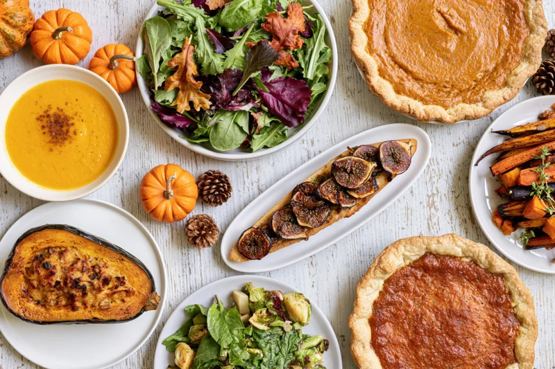 Overhead flat lay of a thanksgiving feast with pumpkin pies, salads, soup, roasted vegetables and autumn decorations on a whitewashed wooden table