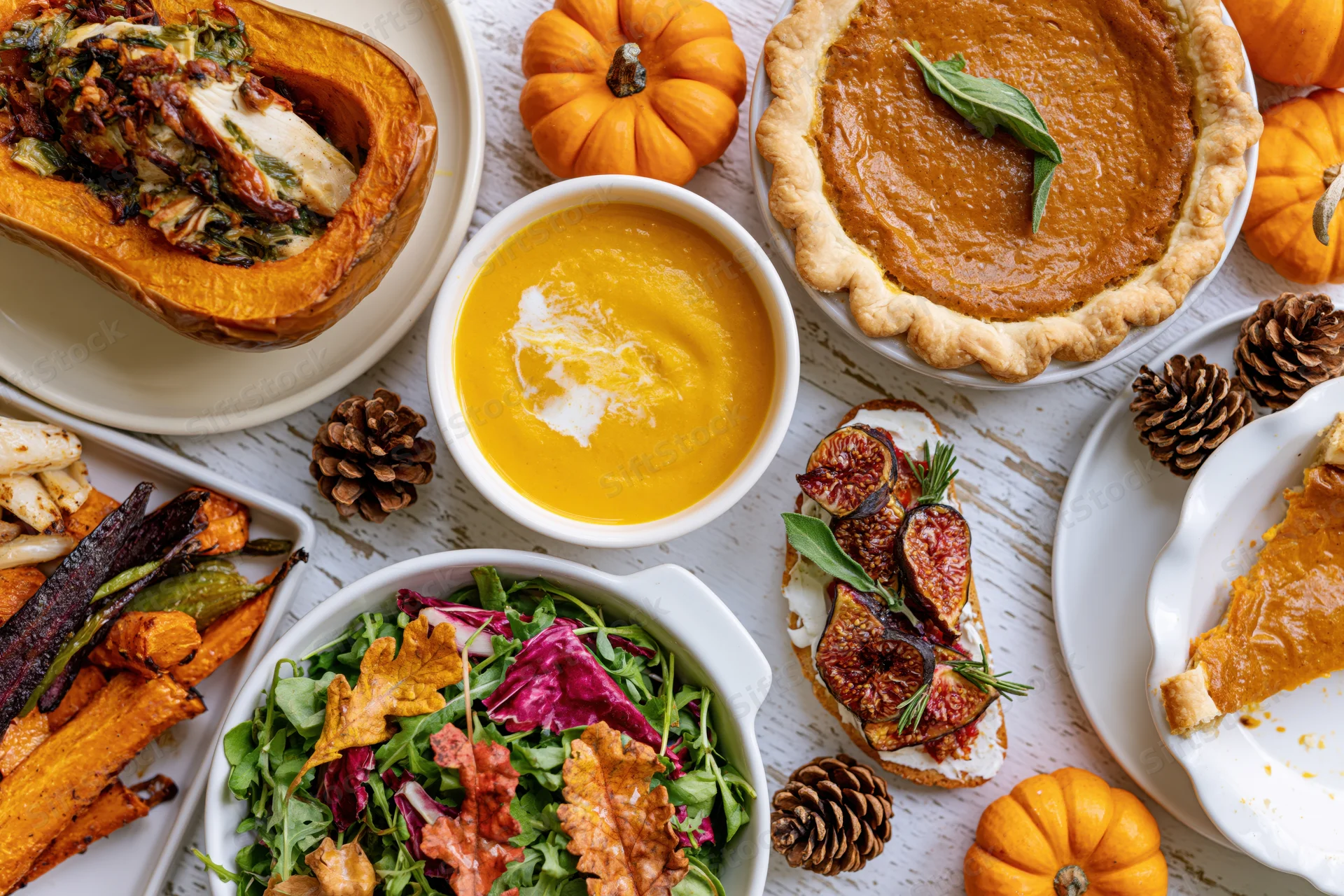 Overhead flat lay of thanksgiving dishes including pumpkin pie, soup, salad, roasted squash and figs on a whitewashed wooden table
