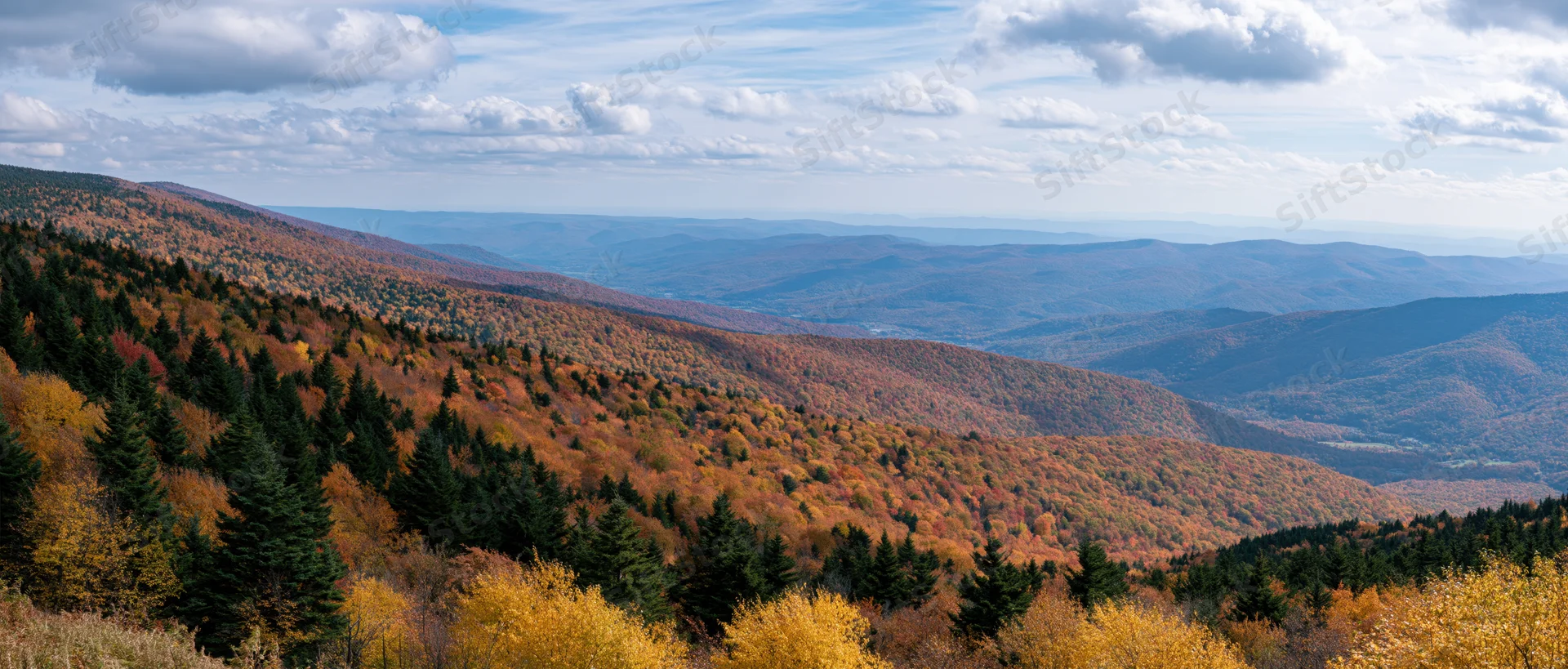 Panoramic view of autumn mountain landscape with mixed conifer and deciduous forest covering rolling hills under a partly cloudy sky