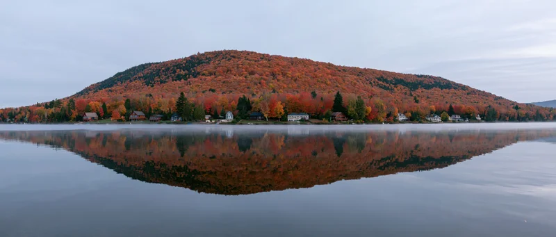 Calm lake reflecting autumn foliage on a forested mountain with homes along the shore