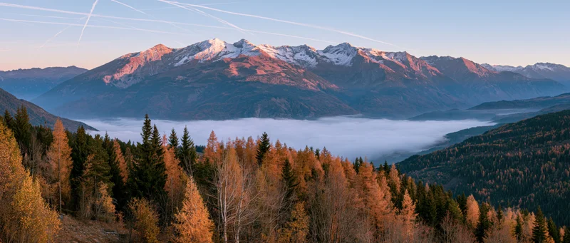 Snow-capped mountain range rising above fog-filled valley with autumn forest in foreground