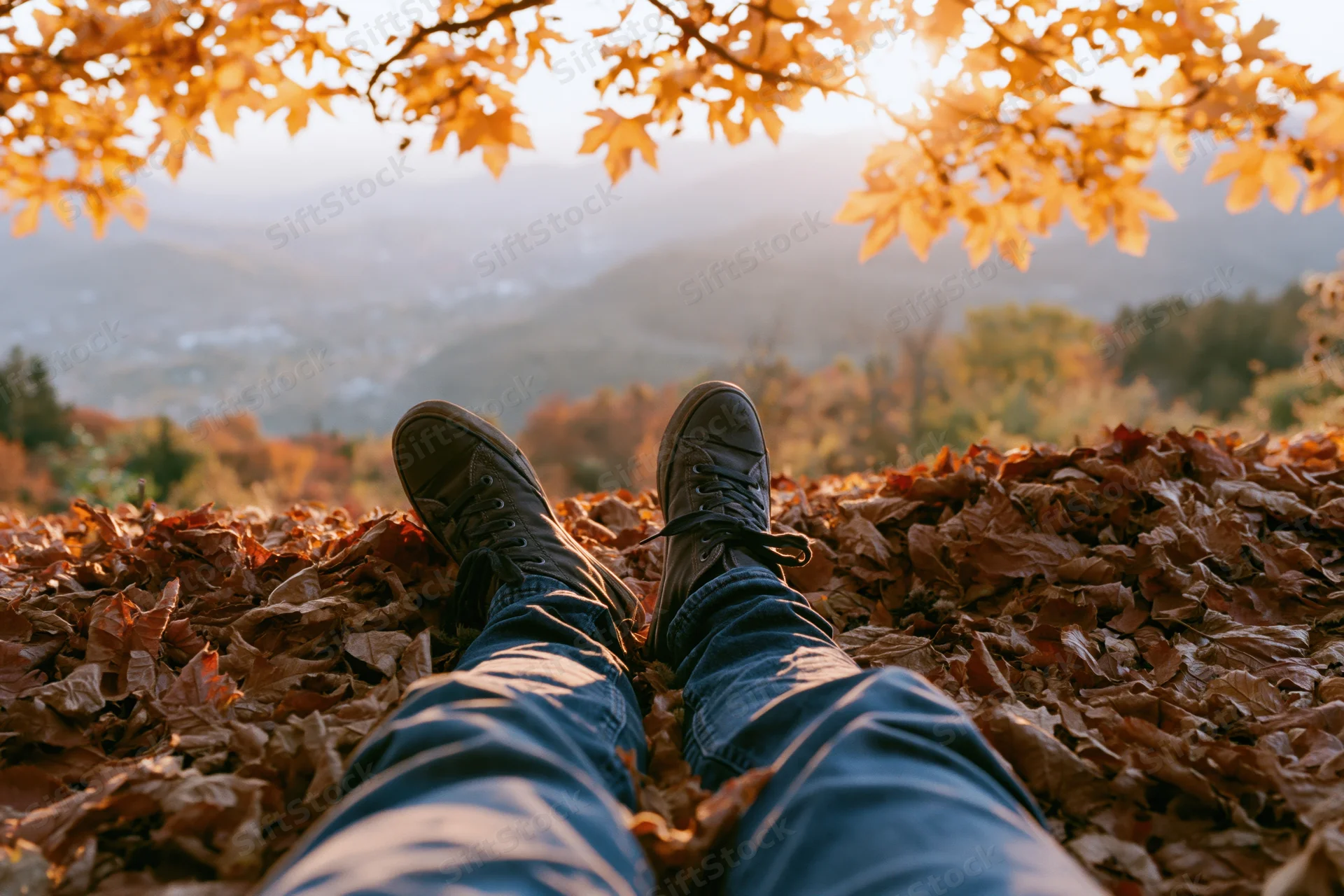 Legs in jeans and sneakers resting on a carpet of fallen autumn leaves overlooking a mountain valley at sunset