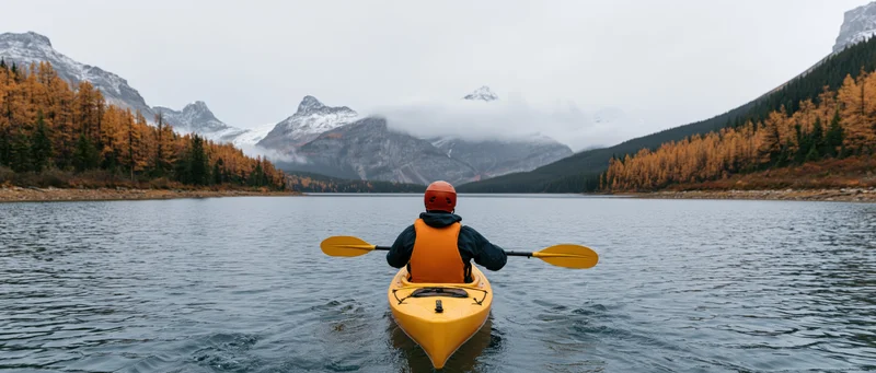 Person paddling yellow kayak on calm lake surrounded by snow-capped mountains and autumn forest