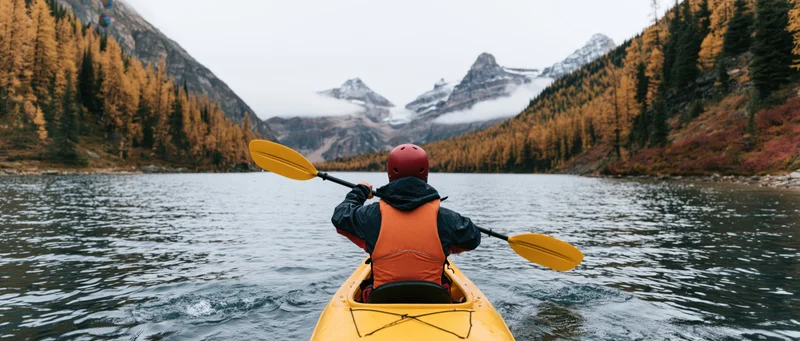 Person paddling yellow kayak on calm alpine lake surrounded by autumn forest and snow-capped mountains