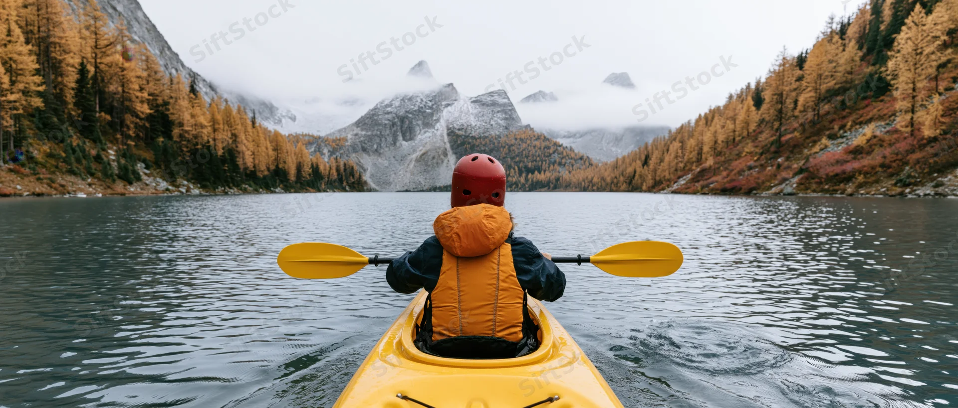 Person paddling yellow kayak on calm alpine lake surrounded by autumn forest and misty mountains