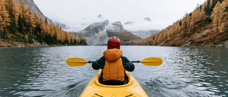 Person paddling yellow kayak on calm alpine lake surrounded by autumn forest and misty mountains