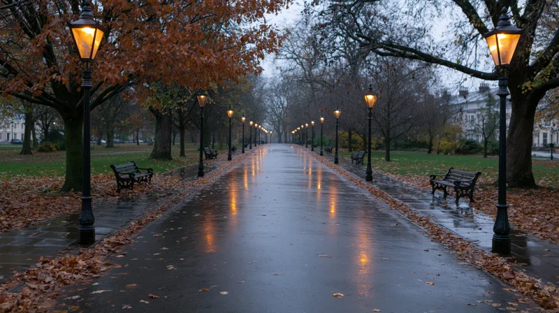 Wet park path lined with glowing lampposts and autumn trees at twilight