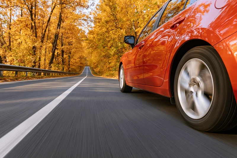Red-orange sedan driving on a two-lane highway through an autumn forest