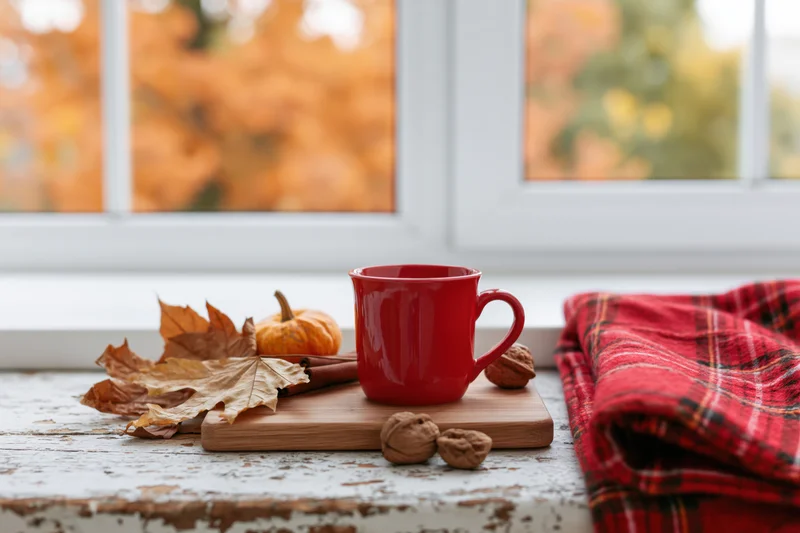 Red ceramic mug on wooden cutting board with autumn leaves and walnuts by window