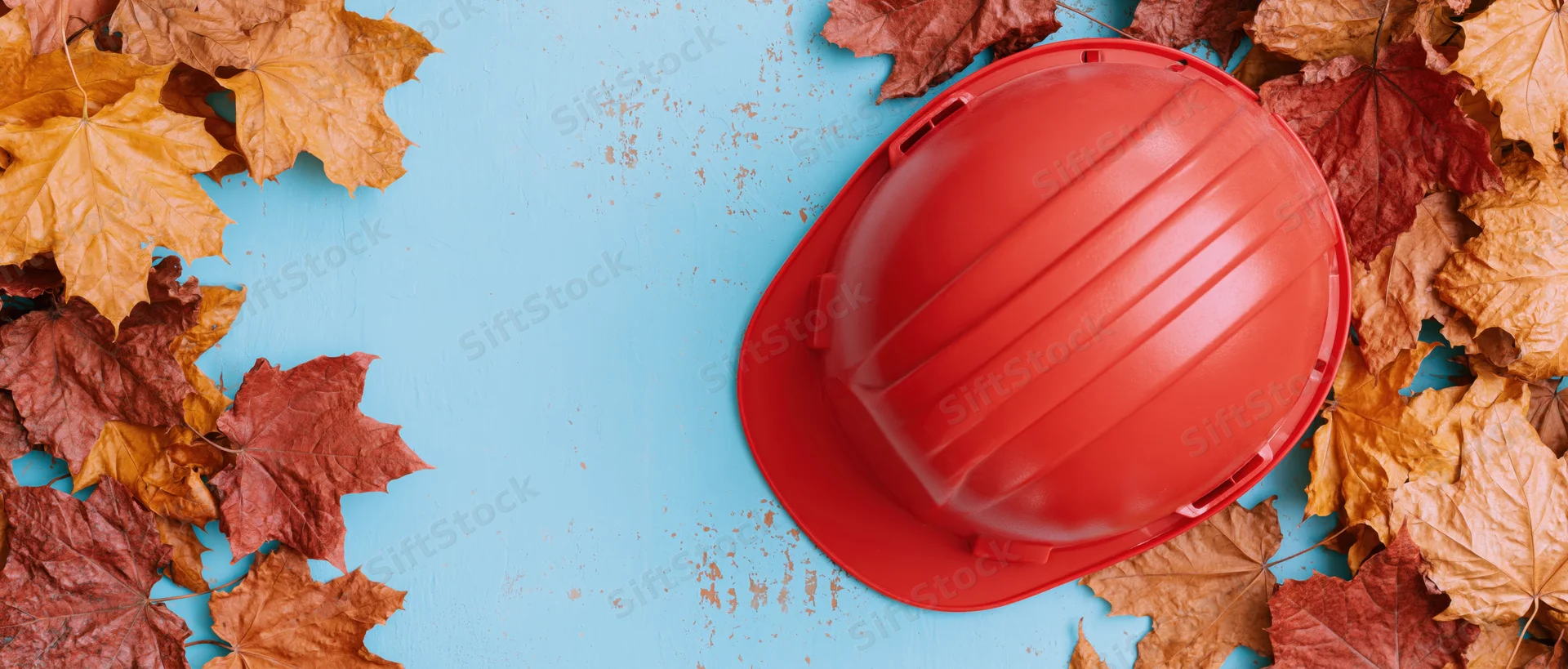 Red construction hard hat lying flat on a worn light blue surface surrounded by autumn maple leaves