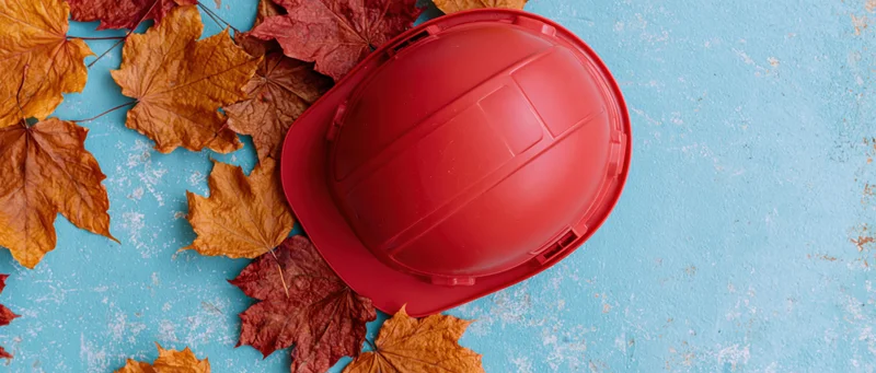 Red construction hard hat resting on a blue textured surface with autumn maple leaves scattered around