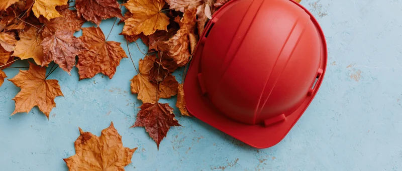Red construction hard hat resting beside dried autumn maple leaves on a worn light blue concrete surface