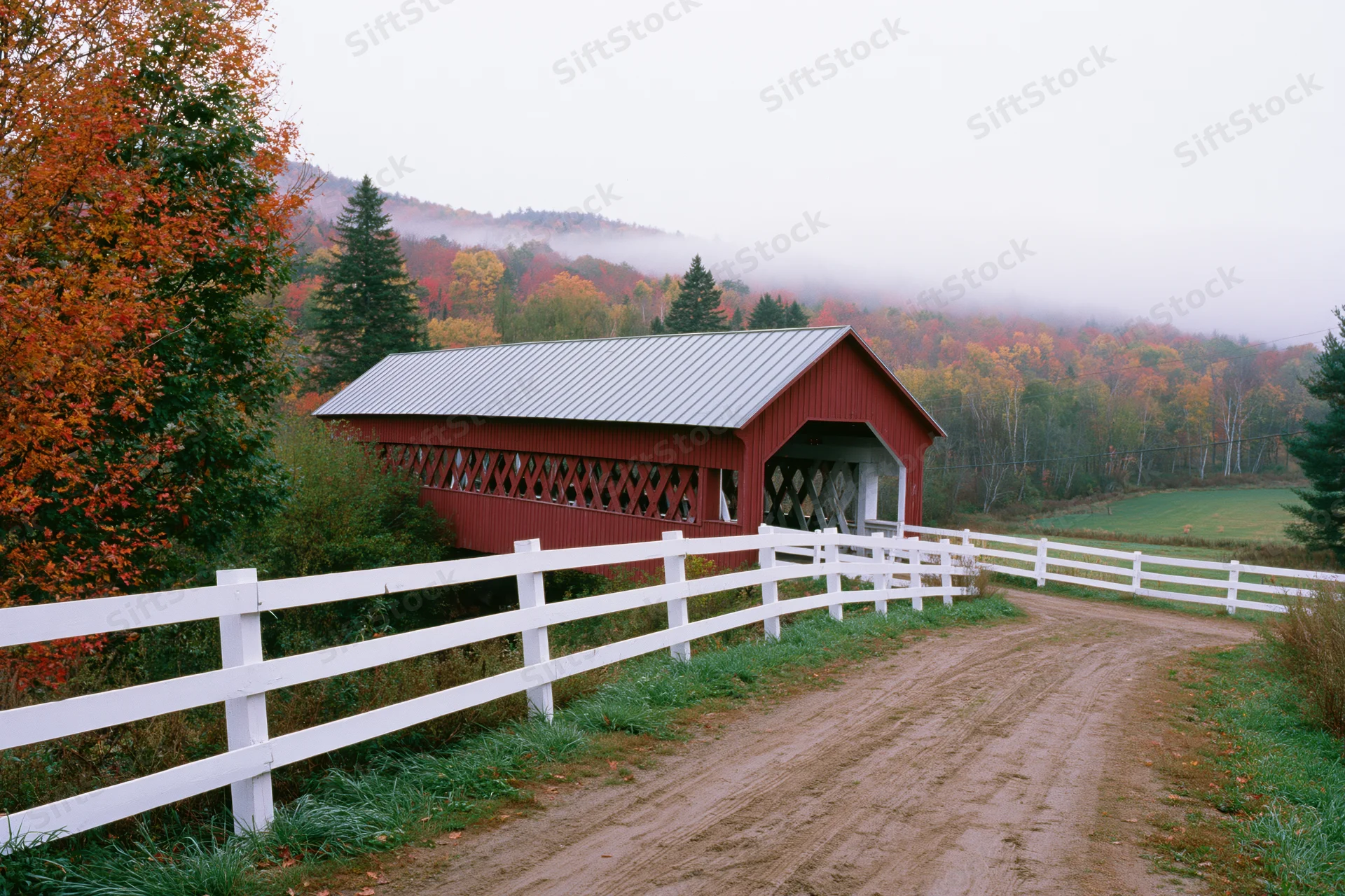 Red covered bridge with metal roof along a dirt road in a foggy rural vermont landscape surrounded by autumn foliage and white fence