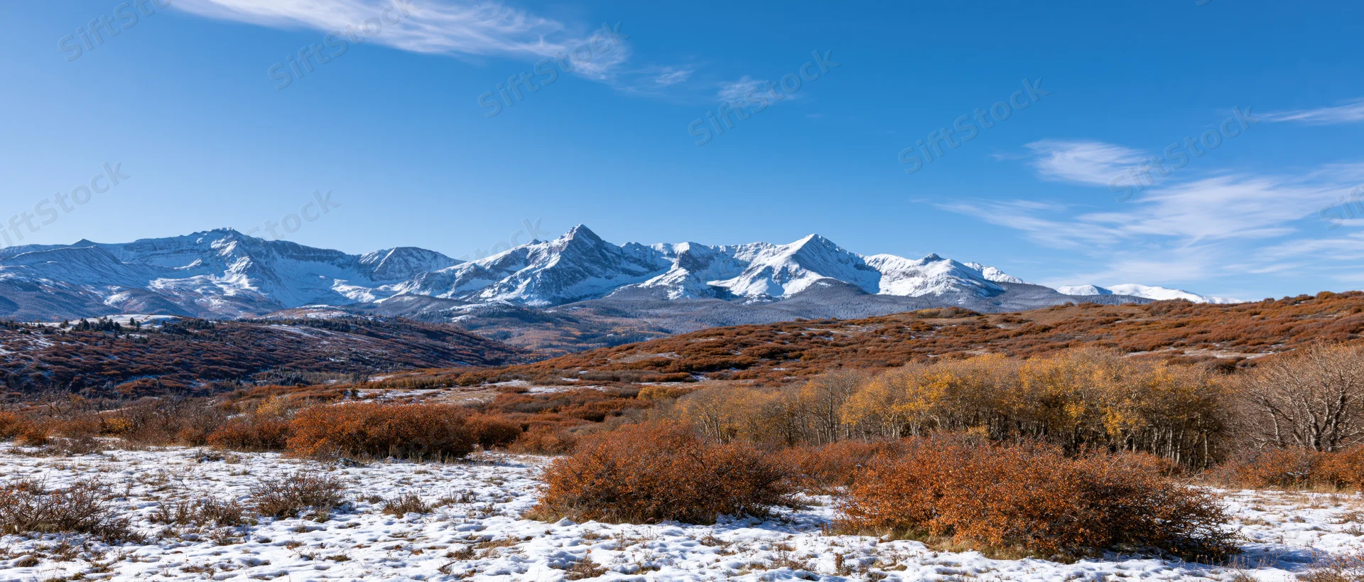 Snow-covered rocky mountains rise behind autumn-colored shrubs in a wide alpine landscape