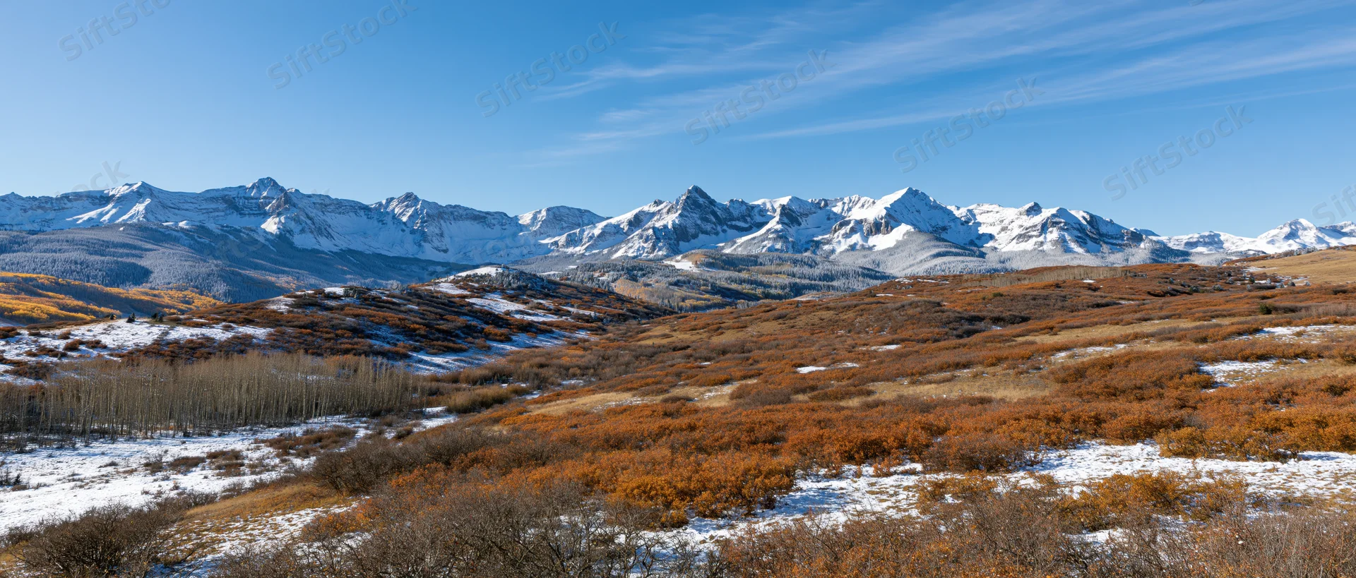 Snow-covered rocky mountains rise above autumn-colored foothills under a clear blue sky