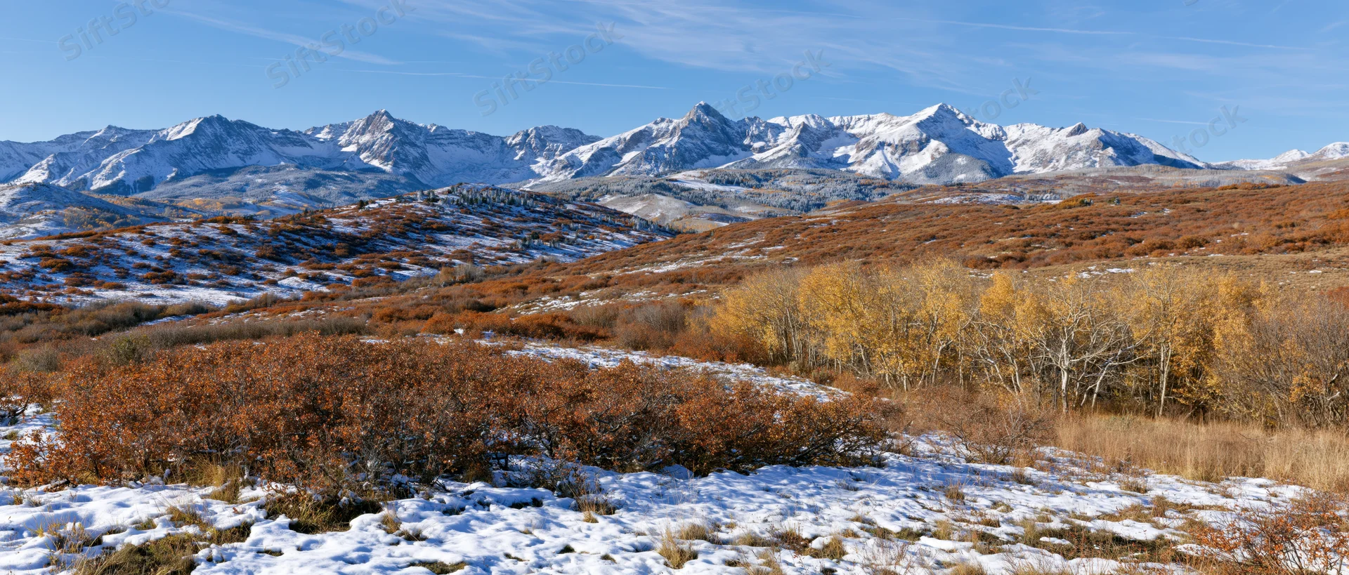 Snow-dusted foothills with autumn-colored aspen trees and rocky mountain peaks under a clear blue sky