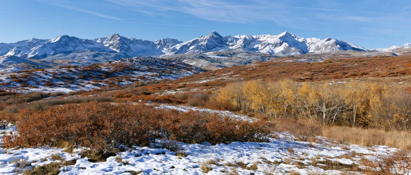 Snow-dusted foothills with autumn-colored aspen trees and rocky mountain peaks under a clear blue sky