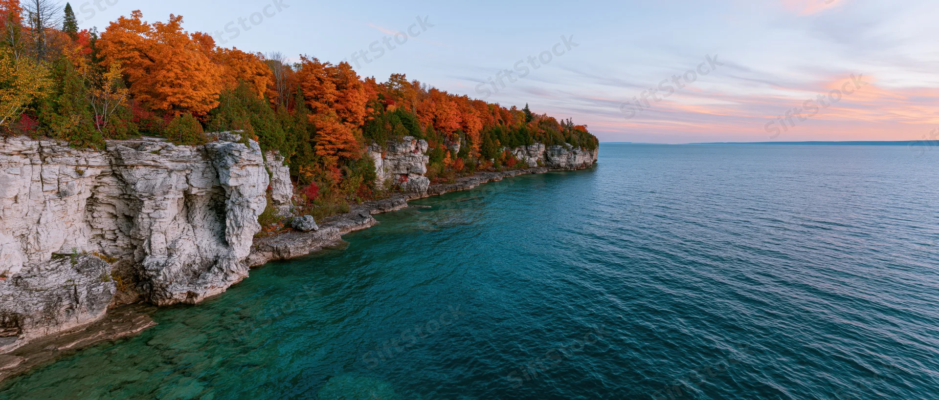 Rocky limestone cliff with autumn trees along a clear turquoise freshwater lake shoreline at sunset