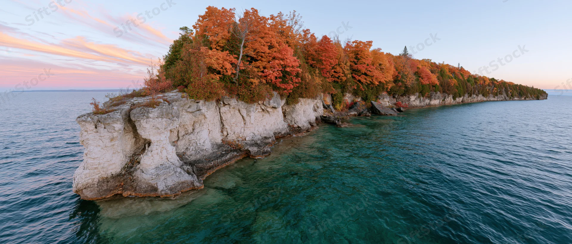Rocky limestone shoreline jutting into clear turquoise freshwater with autumn trees at sunset