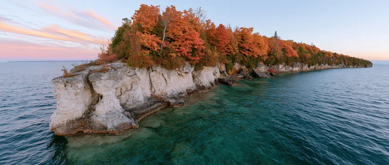 Rocky limestone shoreline jutting into clear turquoise freshwater with autumn trees at sunset