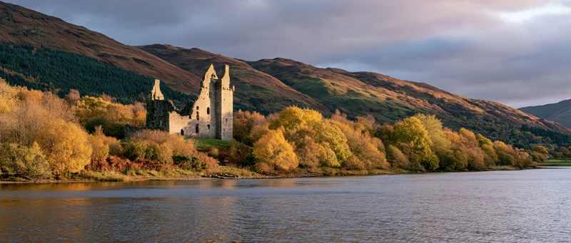 Medieval castle ruins beside a lake surrounded by autumn trees and mountains