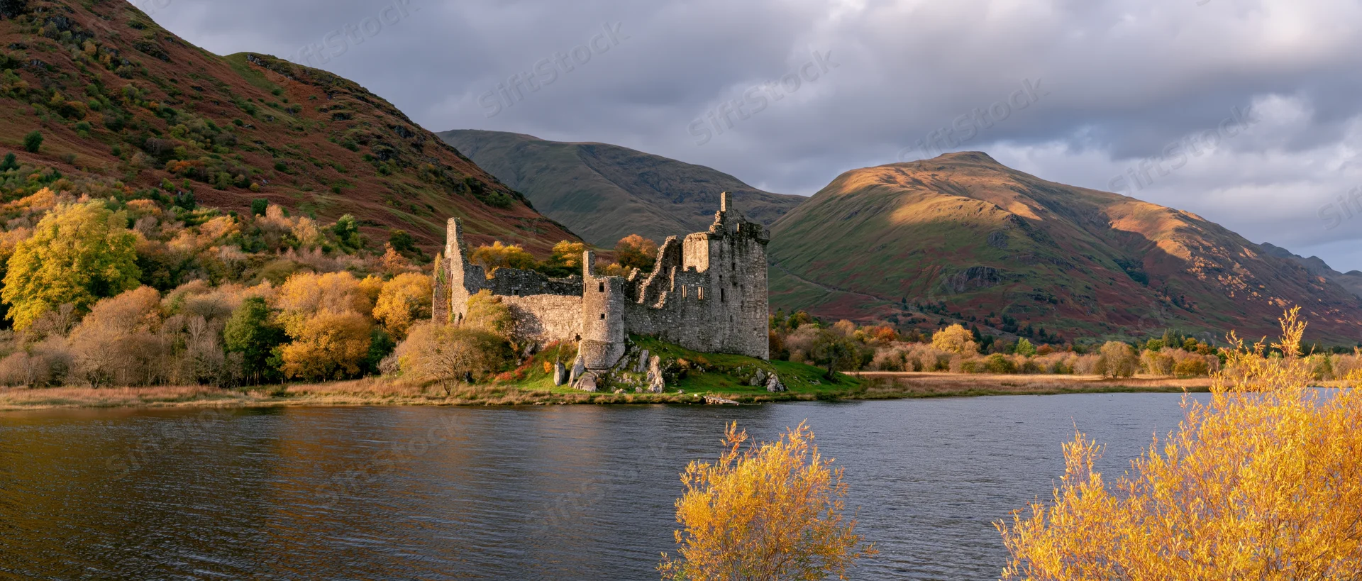 Medieval castle ruins by a lake surrounded by autumn trees and mountains