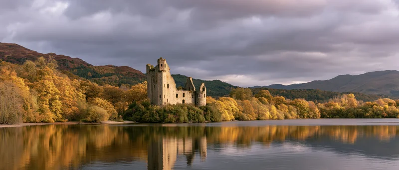 Medieval castle ruins on a lakeshore surrounded by autumn trees and mountains under cloudy sky