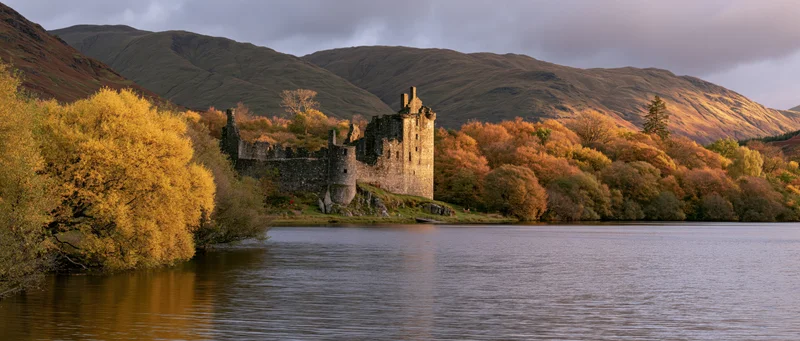 Medieval castle ruins by a lake surrounded by autumn trees and mountains