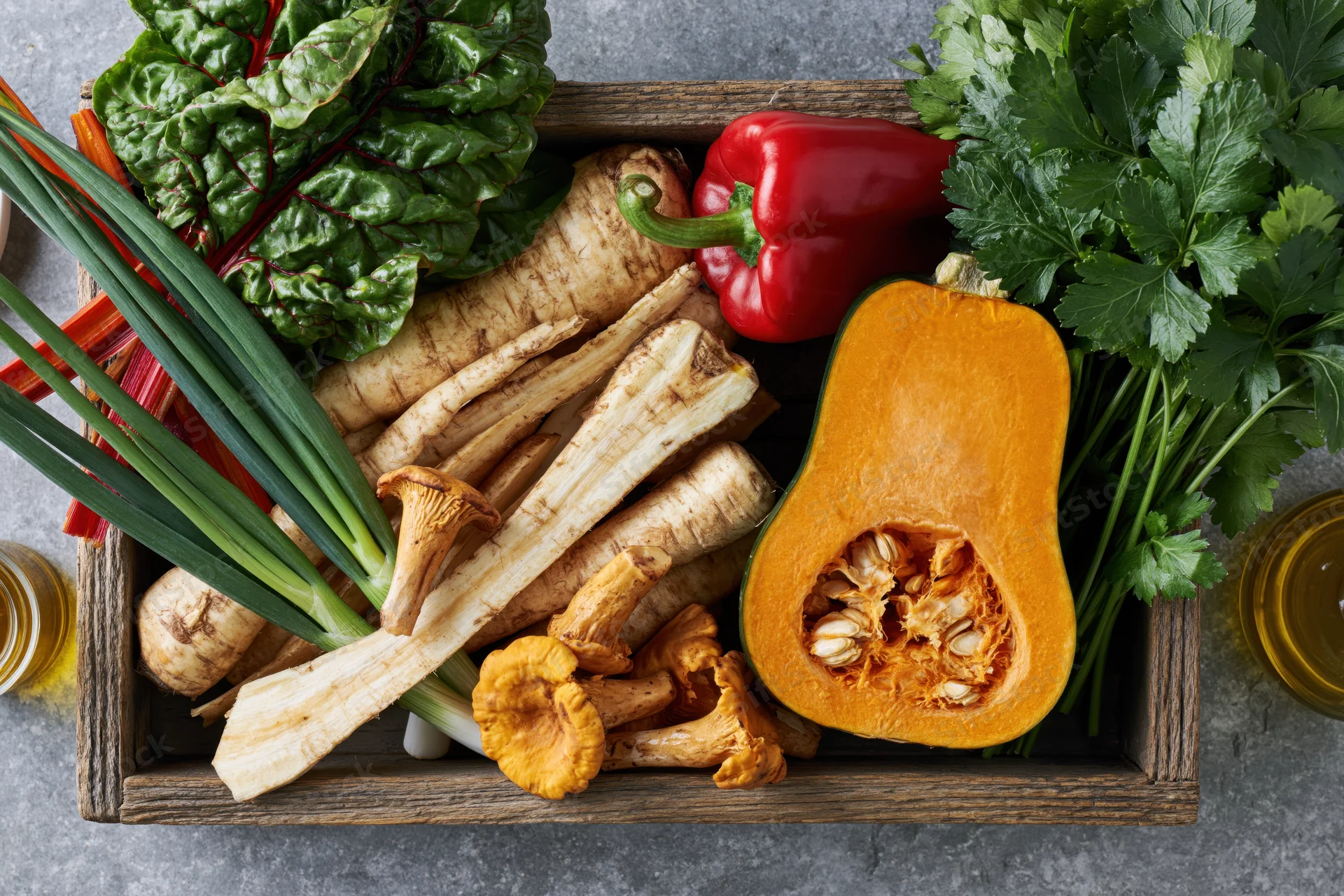 Halved orange pumpkin and parsnips with leafy greens and herbs in a rustic wooden tray on a stone countertop