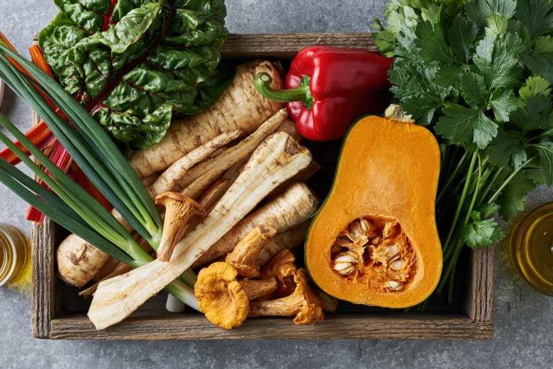 Halved orange pumpkin and parsnips with leafy greens and herbs in a rustic wooden tray on a stone countertop