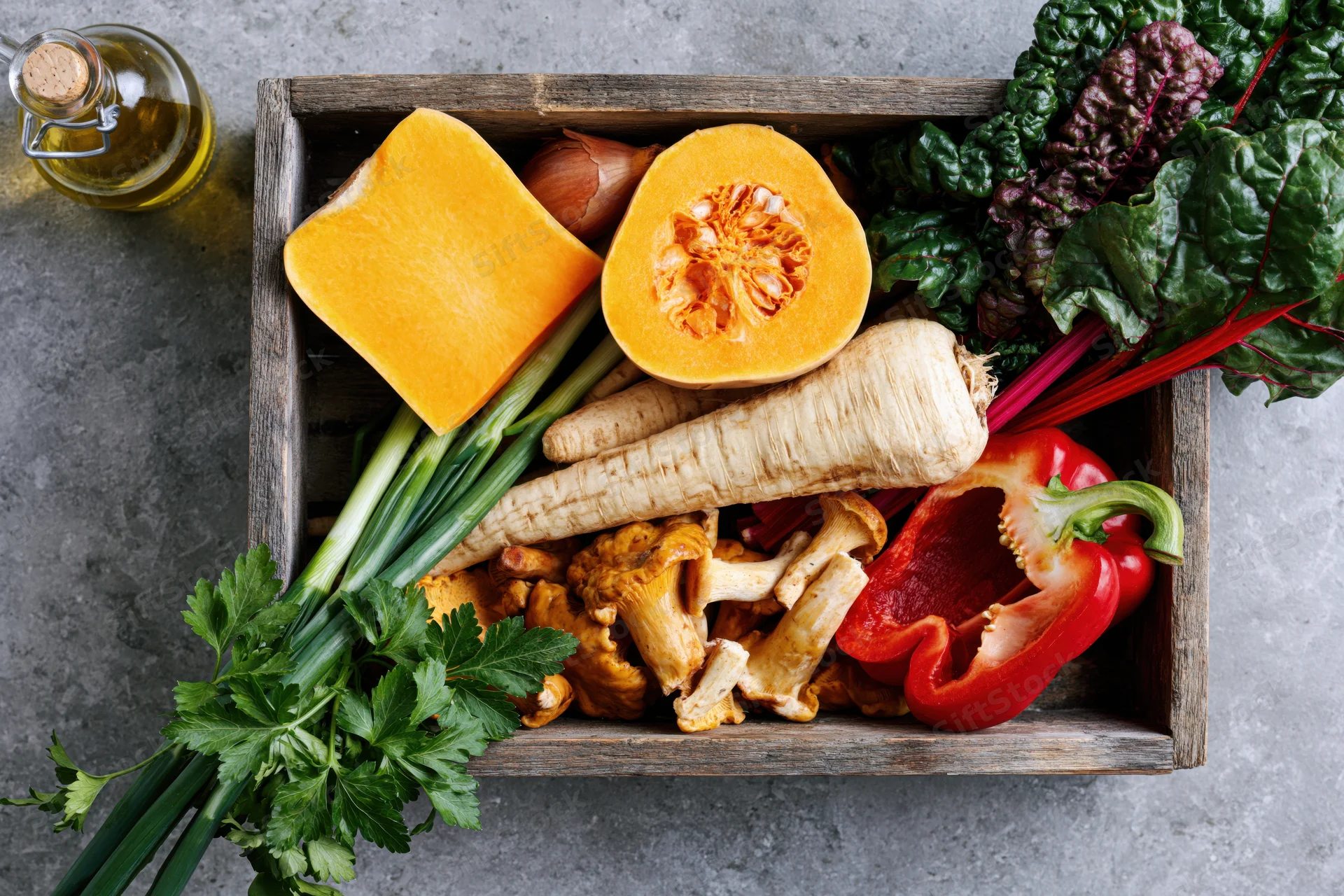 Rustic wooden tray filled with fresh autumn vegetables including pumpkin, parsnips, bell pepper, and herbs on a gray countertop