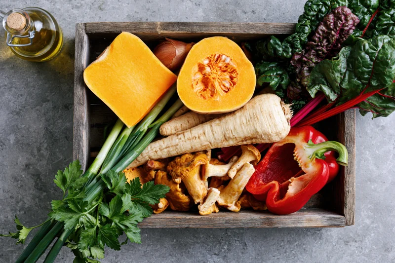 Rustic wooden tray filled with fresh autumn vegetables including pumpkin, parsnips, bell pepper, and herbs on a gray countertop