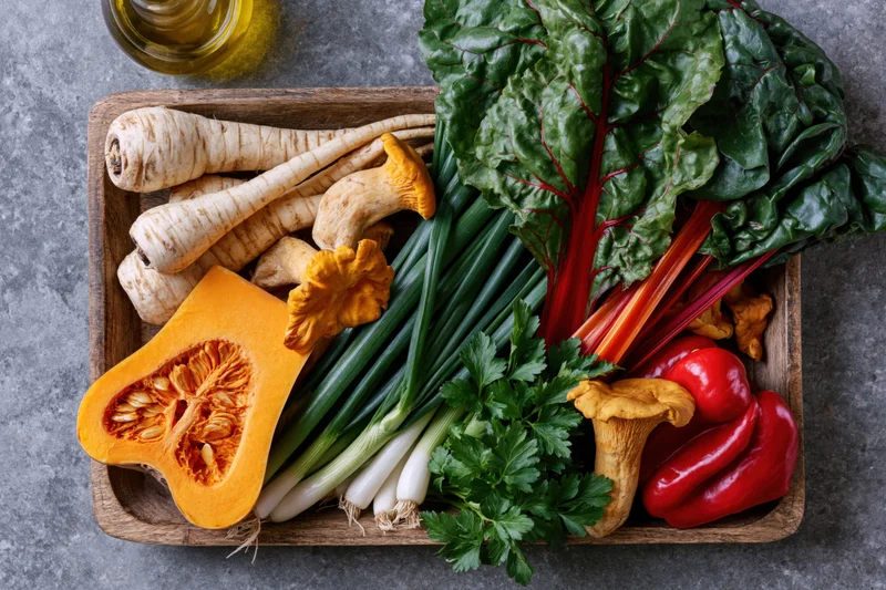 Wooden tray holding pumpkin, parsnips, mushrooms, chard, scallions, peppers and parsley on gray surface