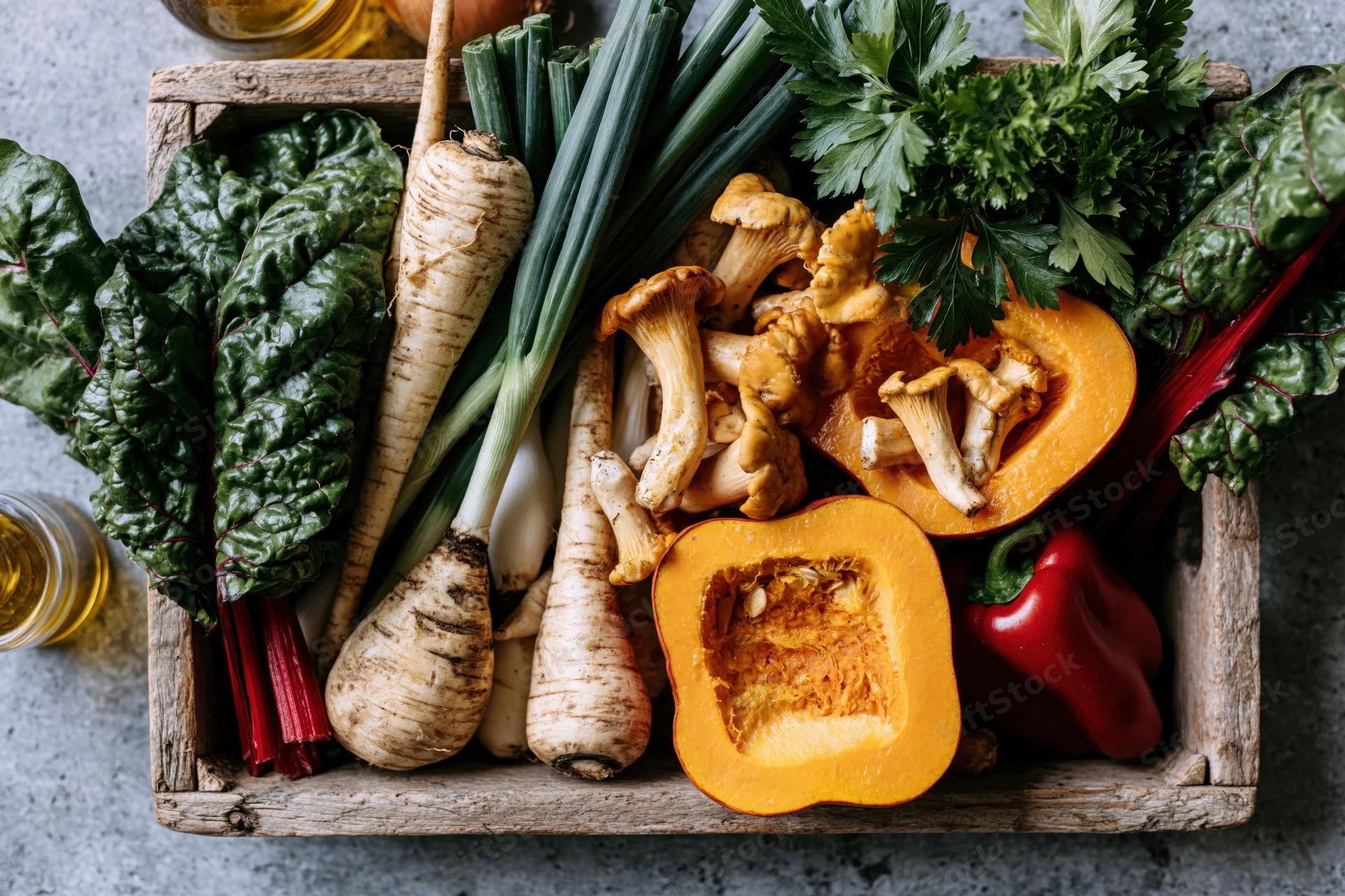 Rustic wooden tray filled with halved pumpkin, parsnips, mushrooms, chard, parsley, and bell pepper on gray surface