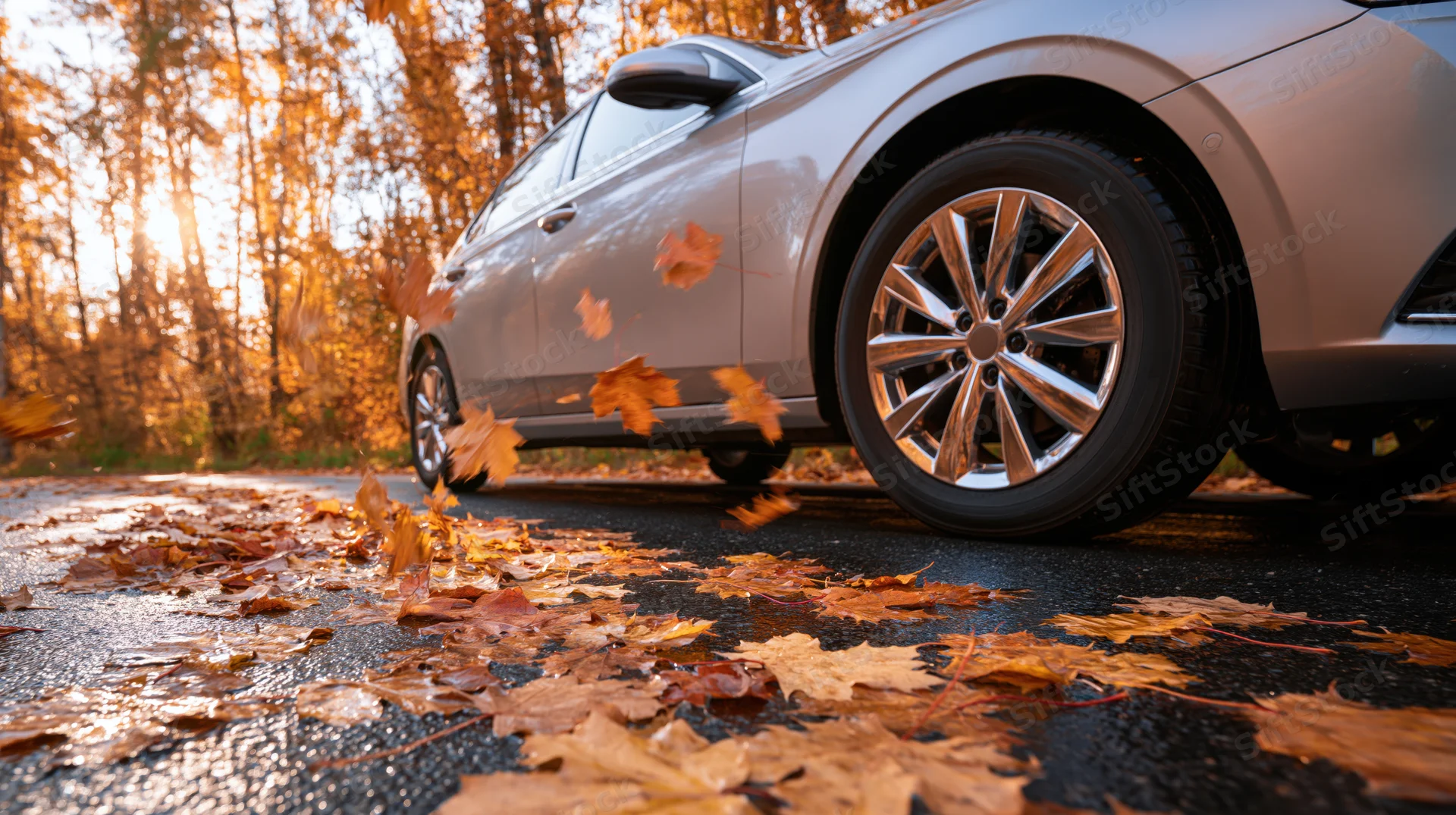 Silver sedan driving on wet autumn road with swirling leaves around wheels